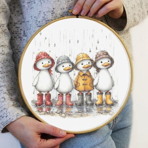 Pode incluir: Um bordado em ponto cruz num bastidor de madeira apresenta quatro patinhos com roupa de chuva. Cada patinho usa um chapéu e botas de chuva de cores diferentes: vermelho, cinzento, amarelo e castanho. O fundo mostra a chuva a cair sobre uma superfície reflectora.