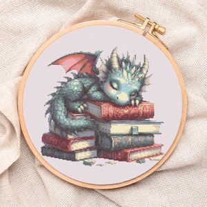 Könnte beinhalten: Kreuzstich-Stickerei eines schlafenden Babydrachens auf einem Bücherstapel. Der Drache ist türkis und blau mit rosa Flügeln und ruht auf roten und beigen Büchern. Die Stickerei befindet sich in einem Holzreifen, auf cremefarbenem Stoff.