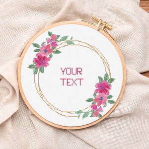 May include: A white embroidery hoop with a gold floral wreath design. The wreath is made of pink flowers and green leaves. The text "YOUR TEXT" is in the center of the hoop.