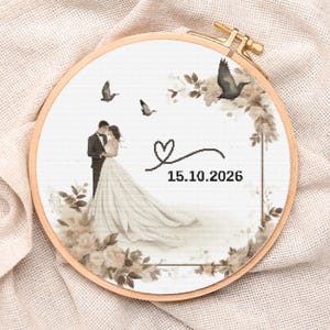 May include: A cross-stitch embroidery hoop featuring a wedding scene. The design includes a couple, doves, floral accents, a heart, and the date 15.10.2026. The embroidery is set in a wooden hoop.