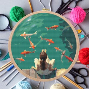 May include: A cross-stitch embroidery hoop with a green and blue background featuring a woman sitting on a wooden dock and watching orange and white fish swim in the water.