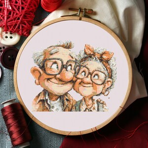 Pode incluir: Um bastidor de bordado em ponto cruz com um casal de idosos sorrindo usando óculos. A obra de arte retrata um homem e uma mulher com cabelos grisalhos, em uma paleta de cores quentes. O bastidor de madeira é cercado por linha vermelha e materiais de costura.