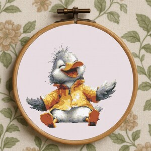 Könnte beinhalten: Kreuzstich-Stickerei einer Cartoon-Ente mit gelber Jacke, die lächelt und die Arme ausstreckt. Die Stickerei befindet sich in einem Holzreifen vor einem floral gemusterten Hintergrund.