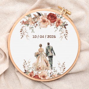 Peut inclure: Un cercle à broder en point de croix avec une scène de mariage. Le motif comprend une mariée et un marié, des éléments floraux rouges, pêche et bruns, et la date 10/04/2026. Le cercle est en bois et possède un fermoir en laiton.