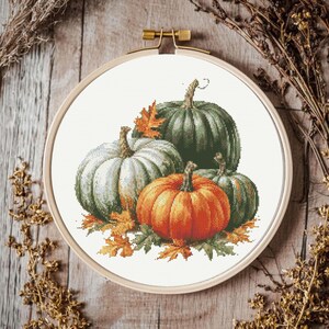 Peut inclure: Un tambour à broder circulaire en bois contenant un motif au point de croix représentant trois citrouilles dans des tons orange, vert sauge et blanc cassé, agrémenté de feuilles d'automne détaillées jaunes et orange sur un fond en tissu blanc.