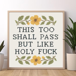 Może przedstawiać: Oprawiona haftem krzyżykowym z napisem "THIS TOO SHALL PASS BUT LIKE HOLY FUCK" w ciemnoniebieskiej nici. Projekt zawiera żółte kwiaty i zielone liście. Rama jest drewniana.