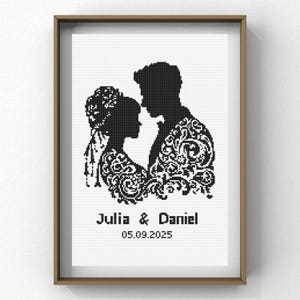 May include: Framed black and white cross-stitch art featuring a couple in silhouette, with the names "Julia & Daniel" and the date "05.09.2025" below. The man's jacket and the woman's dress are decorated with ornate patterns. The frame is a light brown color.
