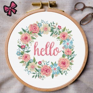 Puede incluir: Un aro de bordado blanco con un diseño de corona floral y la palabra "hello" en letras rosas.