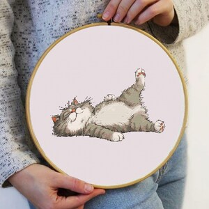 Könnte beinhalten: Ein Stickrahmen mit einem Kreuzstich-Motiv einer entspannten, grau-weißen Katze mit erhobenen Pfoten. Die Katze liegt auf dem Rücken mit geschlossenen Augen. Der Stickrahmen ist vor einem hellrosa Hintergrund platziert.