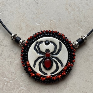 Puede incluir: Un collar de cuentas negras y rojas con un colgante de cerámica blanca que presenta una araña negra con una piedra preciosa roja en el centro.
