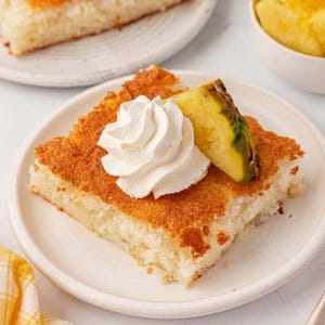 May include: A square slice of golden-brown pineapple cake served on a white plate. The cake is topped with a generous swirl of white whipped cream and a single triangular slice of fresh yellow pineapple, showcasing a moist, textured crumb.