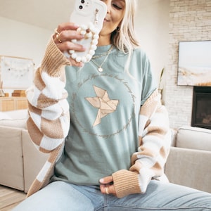 May include: A pale green t-shirt with a paper crane graphic inside a circular text frame. The text reads "The lark flips onto its back, bearing three small perfectly sculpted letters..." The shirt is worn with a beige and white checked cardigan.