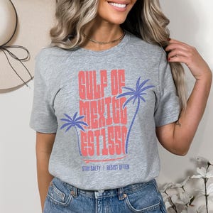 Può includere: T-shirt grigia con la scritta "GULF OF MEXICO EST 1990" in corallo, con due palme blu. La frase "STAY SALTY | RESIST OFTEN" è stampata sotto. Un capo casual per l'uso quotidiano.