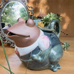 Puede incluir: Tetera de cerámica caprichosa con forma de rana, vestida con un traje con un adorno floral rosa. La tetera tiene una cabeza marrón, ojos negros y un cuerpo azul verdoso. Una pieza decorativa para la cocina o un regalo único.