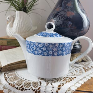 May include: White ceramic teapot with a blue floral band and lid. The teapot has a curved spout and handle. A white swan planter, a dark blue vase with floral designs, and an open book are in the background.