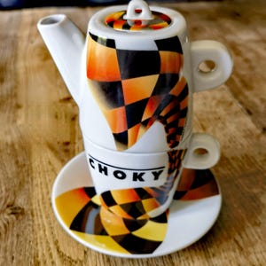 Puede incluir: Un juego de té de cerámica blanca con tetera, taza y platillo. El juego presenta un diseño geométrico audaz en naranja, negro y amarillo. La taza tiene la palabra "CHOKY" impresa.