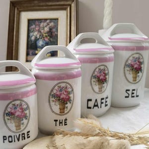 Può includere: Set di quattro barattoli in ceramica bianca con coperchi rosa e motivi floreali. I barattoli sono etichettati con "Poivre", "The", "Cafe" e "Sel".