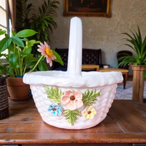Peut inclure: Un panier en céramique blanc avec une anse, décoré de motifs floraux en relief. Le panier présente une texture tissée et est orné de trois fleurs colorées roses, bleues et jaunes, avec des feuilles vertes.