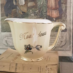 Könnte beinhalten: Cremefarbener Porzellan-Sahnegießer mit goldenem Rand und Griff. Der Sahnegießer trägt den goldenen Schriftzug "Tous deux" und ein Blumendesign. Er steht auf einem Stapel alter Bücher mit französischem Text vor einem Vintage-Druck.