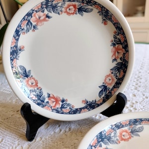 Puede incluir: Plato de cerámica blanca con diseño floral. El plato presenta un borde de hojas azules y flores color coral. El plato se exhibe sobre un soporte negro, mostrando su patrón decorativo. El plato es parte de un juego, perfecto para servir comidas.