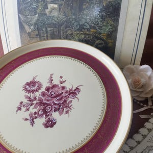 May include: A cream-coloured decorative plate with a burgundy border and gold trim. The plate features a floral design in shades of burgundy. The background includes a framed botanical print and a fabric rose.