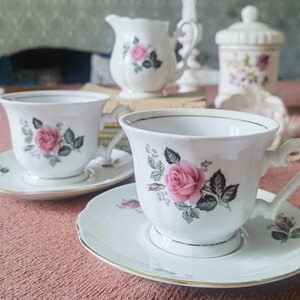May include: A white porcelain tea set featuring teacups, saucers, a creamer, and a lidded jar. Each piece is decorated with pink roses, green leaves, and gold trim. The teacups have decorative handles.