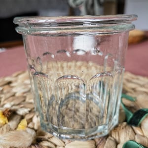 Peut inclure: Un pot en verre transparent de style vintage, avec un large bord et un motif festonné décoratif sur les côtés. Le pot est posé sur un dessous de plat tissé de tons naturels. Le verre est transparent.