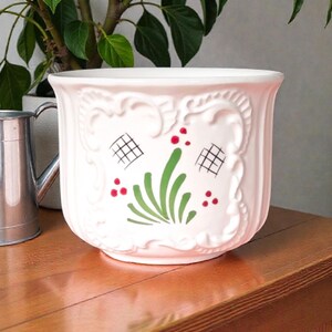 Peut inclure: Un pot de fleurs en céramique rose clair avec un motif floral décoratif. Le pot a un bord blanc et est orné d'accents peints en vert, rouge et noir. Le pot est posé sur une surface en bois.