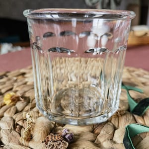 Peut inclure: Un verre transparent à facettes avec un bord arrondi. Le verre présente des lignes verticales et de petites indentations ovales. Le verre est posé sur un tapis tissé brun clair avec des fleurs séchées et des rubans verts.