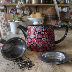 May include: A black teapot with a floral pattern in red, pink, and white, with a black handle and spout. A stainless steel tea infuser and loose tea leaves are next to the teapot. The teapot is on a wooden surface.