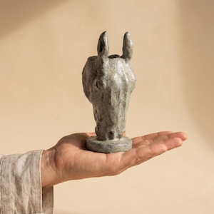 May include: A small, hand-sculpted ceramic horse head figurine with a textured, mottled grey-green glaze. The sculpture features upright ears, a defined snout, and a circular base, showcasing a rustic, uneven surface finish throughout the piece.
