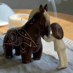Peut inclure: Figurine de cheval en céramique marron foncé avec des marques décoratives blanches et une crinière crème. Une petite figurine crème d'une personne touche la tête du cheval. Une bougie allumée et de la fumée sont en arrière-plan.
