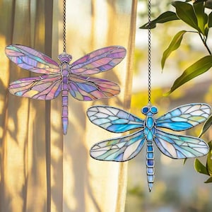 May include: Two stained glass dragonfly ornaments hanging from chains. One dragonfly features purple, pink, and yellow wings, while the other has blue and white wings. The dragonfly bodies are outlined in a dark metal.