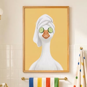 May include: A framed wall art print featuring a white goose with a white towel wrapped around its head and two green cucumber slices over its eyes, set against a solid yellow background. The goose has an orange beak and a long white neck.