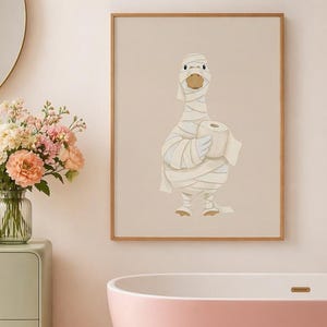 May include: A framed wall art print featuring a cartoon goose wrapped in white bandages like a mummy, holding a roll of toilet paper. The illustration is set against a solid beige background, presented in a simple light-colored wooden frame.