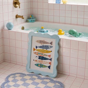 May include: A framed art print featuring colorful fish illustrations in a light blue scalloped frame. The artwork is displayed in a bathroom setting with a blue and white checkered rug, and toy boats and animals in the bathtub.