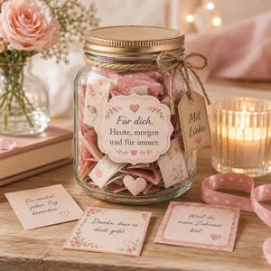Può includere: Un barattolo di vetro trasparente con coperchio dorato riempito di piccoli foglietti piegati con motivi floreali rosa. Un'etichetta riporta: Für dich. Heute, morgen und für immer. Un cartellino marrone legato con spago riporta: Mit Liebe. Tre note sono davanti.