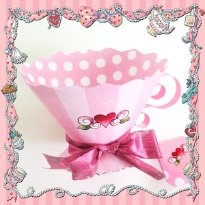 May include: A pink paper teacup with a scalloped rim and a pink bow. The teacup has a striped design with white polka dots and a heart design. The image is framed with a pink border with hearts and pearls.