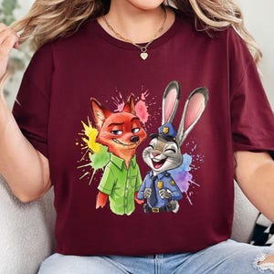 May include: Maroon t-shirt featuring a colorful graphic of Nick Wilde the fox and Judy Hopps the rabbit from Zootopia. Nick is wearing a green shirt, and Judy is in a police uniform. The word "MAROON" is printed at the bottom.
