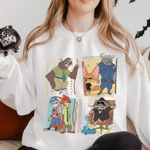 Puede incluir: Sudadera blanca con un colorido gr&aacute;fico de dibujos animados de personajes de una pel&iacute;cula de animaci&oacute;n. El gr&aacute;fico incluye un perezoso, un zorro, un b&uacute;falo y otros personajes. La palabra "WHITE" est&aacute; impresa en la parte inferior.