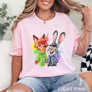 May include: Light pink t-shirt featuring a colorful graphic of Nick Wilde and Judy Hopps from Zootopia. Nick is wearing a green shirt, and Judy is in a police uniform. The text "LIGHT PINK" is at the bottom.