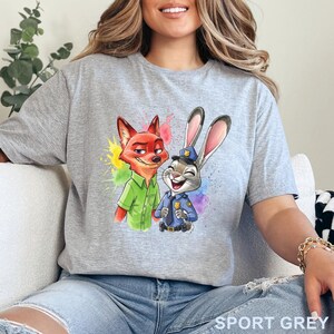 May include: Sport grey t-shirt featuring a colorful graphic of Nick Wilde and Judy Hopps from Zootopia. Nick is a fox in a green shirt, and Judy is a rabbit in a police uniform. The shirt has a watercolor splash effect.