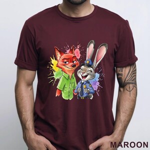 May include: Maroon t-shirt featuring a colorful graphic of a fox and a rabbit from Zootopia. The fox is wearing a green shirt, and the rabbit is in a police uniform. The design includes colorful paint splatters.