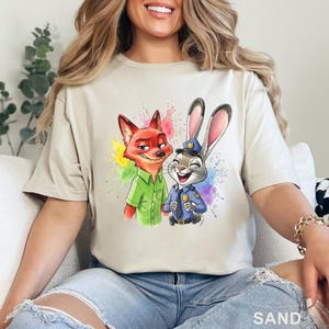 May include: Sand-colored t-shirt featuring a colorful graphic of a fox and a rabbit, with paint splatter accents. The fox is wearing a green shirt, and the rabbit is in a police uniform. The word "SAND" is printed at the bottom.