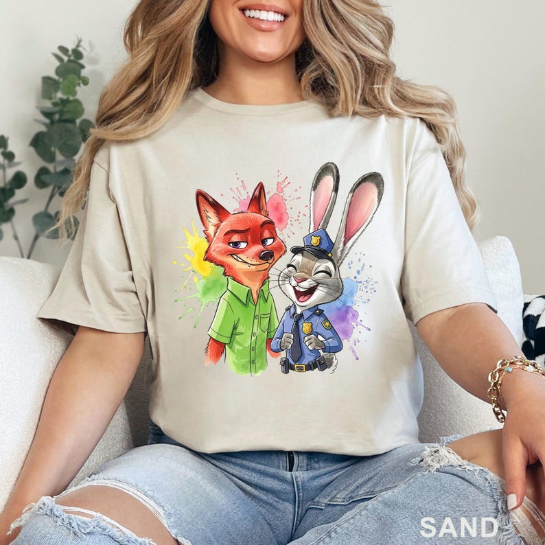 May include: Sand-colored t-shirt featuring a colorful graphic of a fox and a rabbit, with paint splatter accents. The fox is wearing a green shirt, and the rabbit is in a police uniform. The word "SAND" is printed at the bottom.