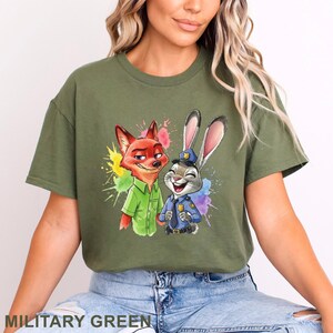 May include: Military green t-shirt featuring a colorful graphic of a fox in a green shirt and a rabbit in a police uniform. The graphic has a watercolor paint splash effect. The text "MILITARY GREEN" is at the bottom.