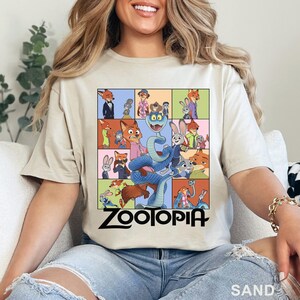 Peut inclure: T-shirt couleur sable avec un motif color&eacute; de personnages de Zootopie, dont Nick Wilde et Judy Hopps. Un grand serpent bleu est au centre. Le mot "ZOOTOPIA" est imprim&eacute; en noir en dessous.
