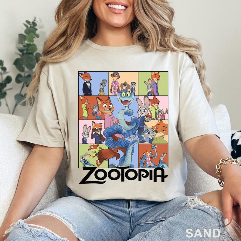 Peut inclure: T-shirt couleur sable avec un motif color&eacute; de personnages de Zootopie, dont Nick Wilde et Judy Hopps. Un grand serpent bleu est au centre. Le mot "ZOOTOPIA" est imprim&eacute; en noir en dessous.