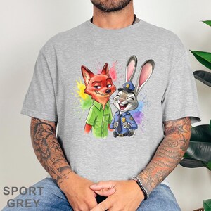 May include: Sport grey t-shirt featuring a colorful illustration of Nick Wilde the fox and Judy Hopps the rabbit from Zootopia. Nick is wearing a green shirt, and Judy is in a police uniform. The design includes colorful paint splatters.