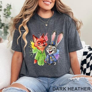 May include: Dark heather gray t-shirt featuring a colorful graphic of Nick Wilde and Judy Hopps from Zootopia. Nick is wearing a green shirt, and Judy is in a police uniform. The text "DARK HEATHER" is at the bottom.